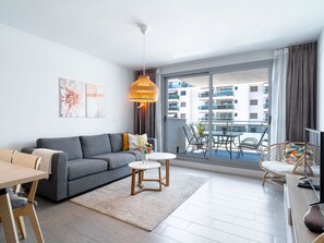 Un salon moderne avec un canapé gris, une table basse et un coin repas comprenant une table et des chaises. Une grande fenêtre offre une vue sur les bâtiments environnants.