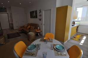 A modern living room with a sofa, dining table set for two, and a kitchen area with yellow cabinets. - Romeo House (Cagliari)