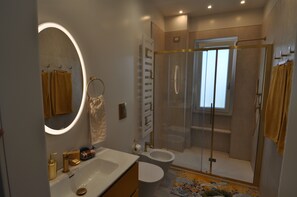 Une salle de bain moderne avec une douche à l’italienne en verre, un grand miroir rond avec éclairage intégré et un lavabo blanc agrémenté de robinetterie dorée.