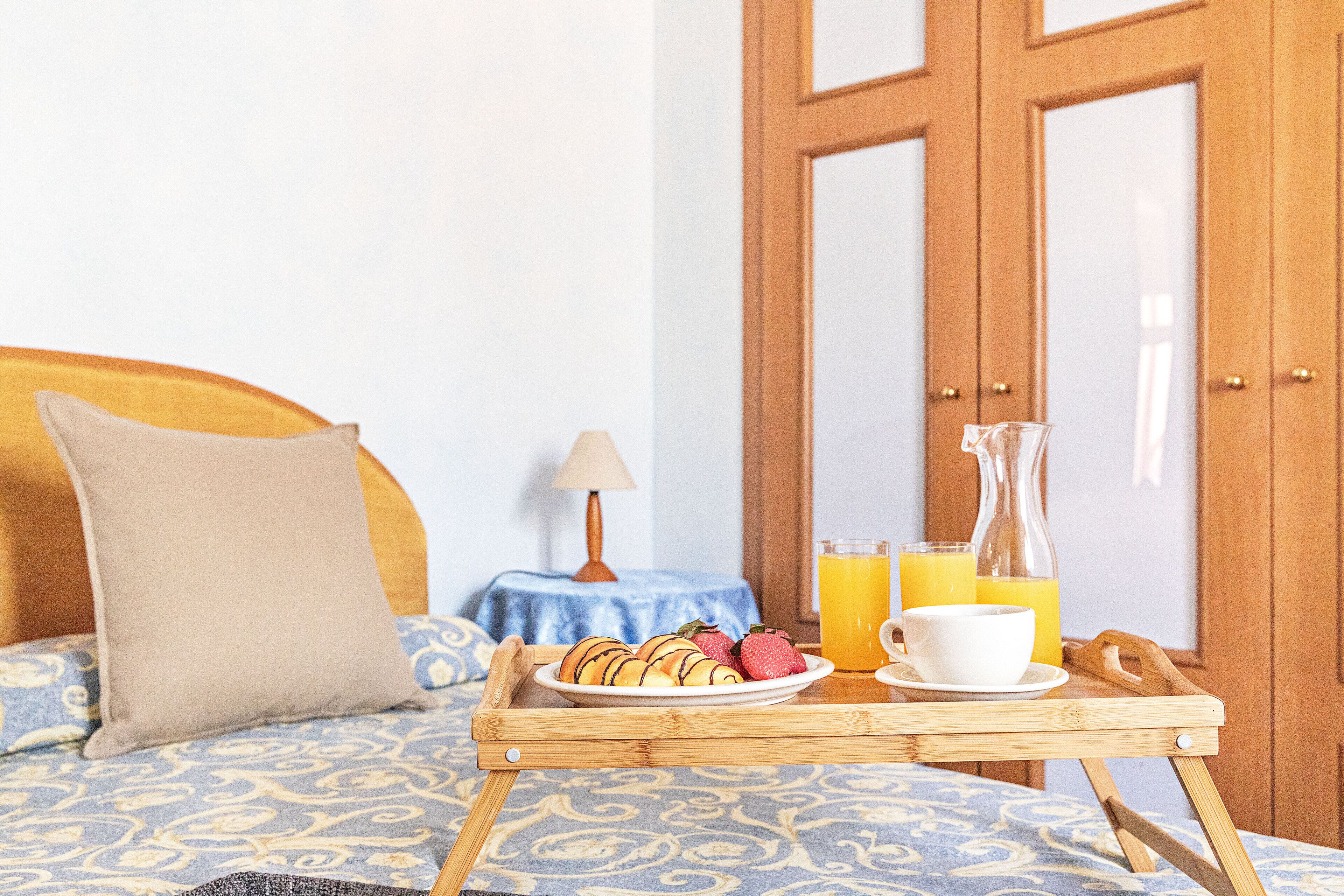 A bed with a headboard, a bedside table with a lamp, a tray with pastries and a glass of orange juice, and a wooden door in the background.