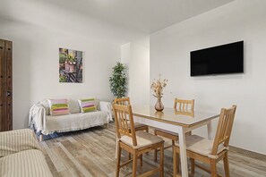 A dining area with a glass-top table, wooden chairs, a sofa with striped cushions, a potted plant, and a flat-screen TV mounted on the wall. - Holiday Home 'El Islote del Sur' with Private Pool, Wi-Fi & Air Conditioning (Conil de la Frontera)