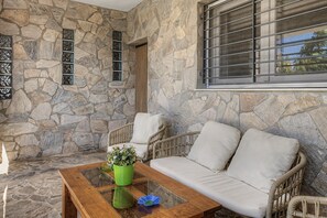 Ein Zimmer mit Steinwänden, das eine Sitzecke mit Sofa und Sesseln, ein Fenster mit Gitterstäben sowie einen Glastisch mit Blumenvase beinhaltet.