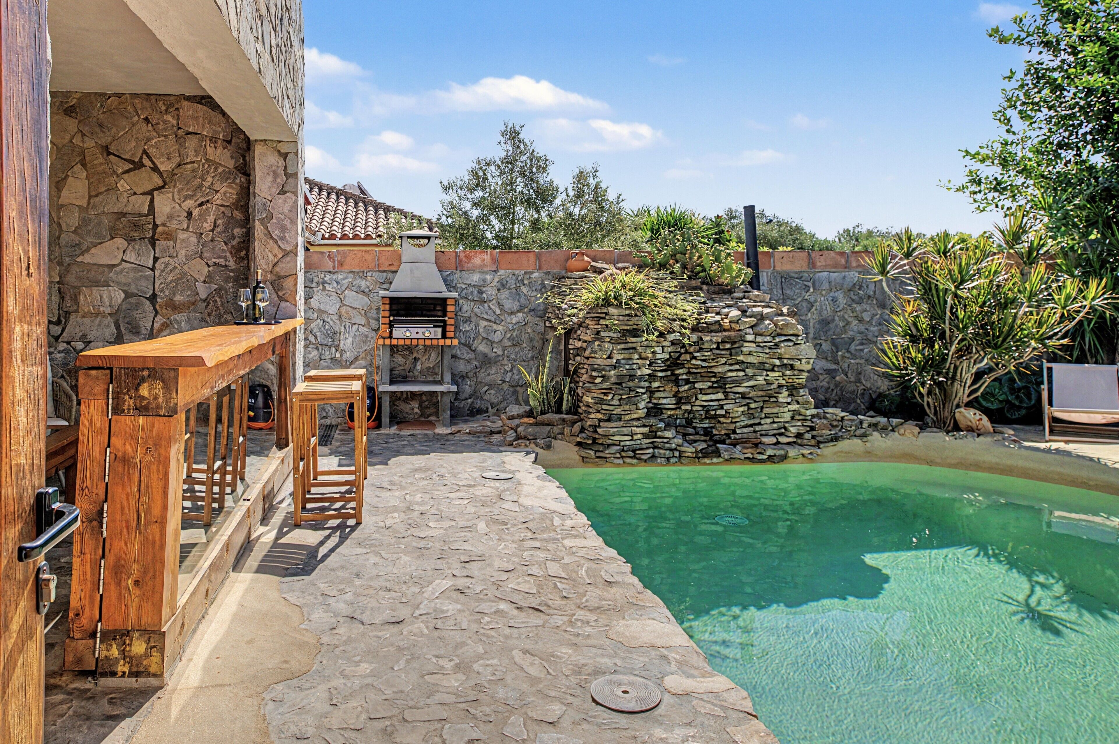 A stone patio with a pool, a wooden bar, and a stone wall with plants.