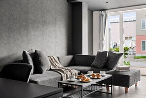 A modern living room with a grey sofa, a coffee table with pastries, and a view of the outside. - HT-I (Asahikawa)