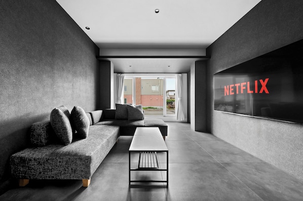 A modern living room with a sectional sofa, a coffee table, and a large window with a view of buildings. - HT-I (Asahikawa)