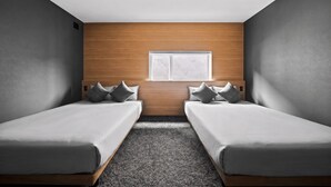 A room with two single beds, a wooden accent wall, a small window, and a textured carpet. - HT-I (Asahikawa)
