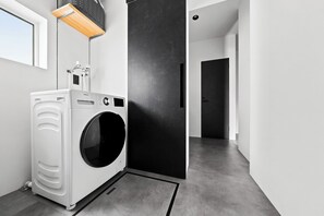 A modern laundry room with a front-loading washer and dryer, a black door, and a small window. - HT-I (Asahikawa)