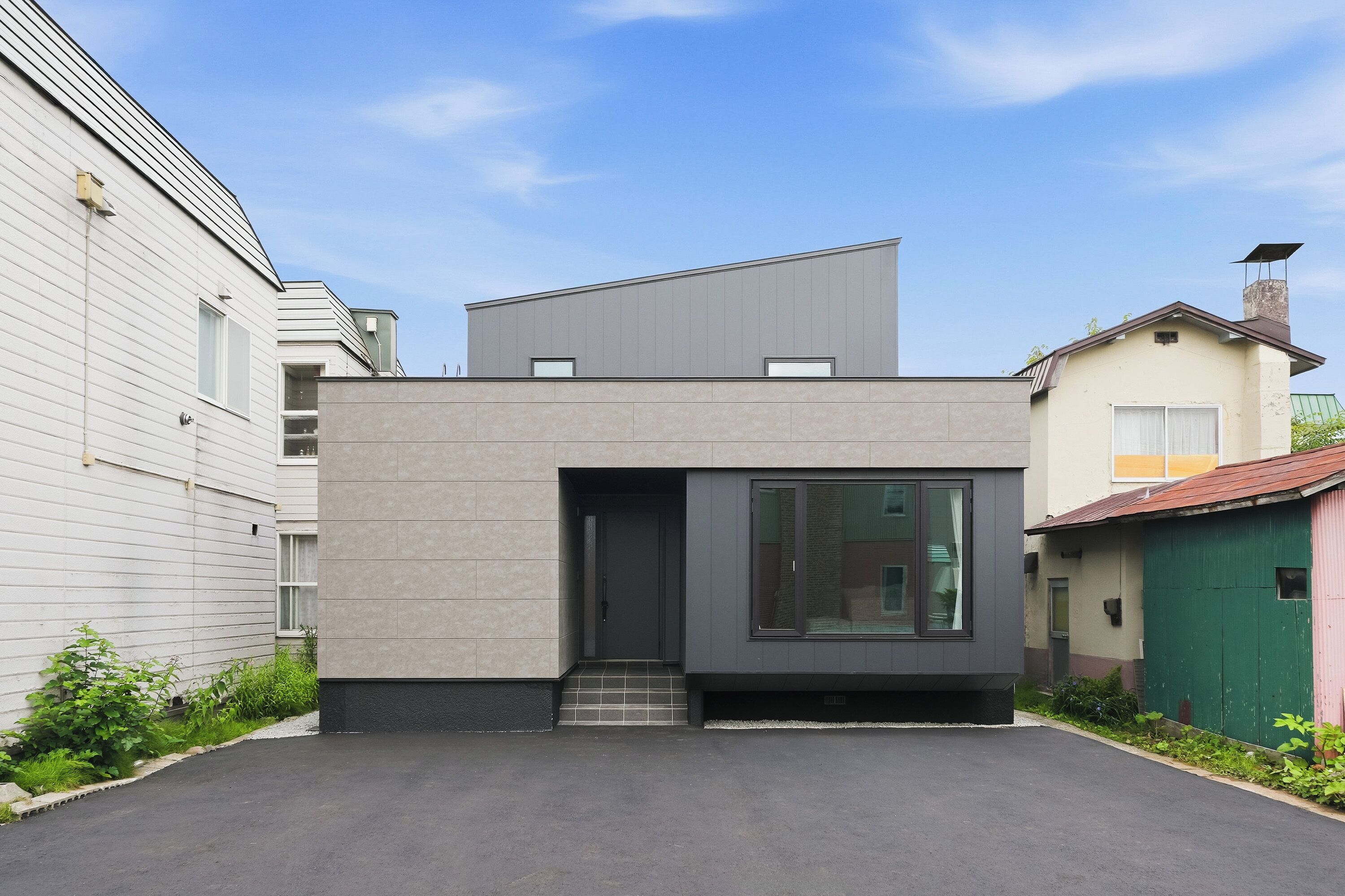Une maison moderne à deux étages, dotée d’un toit plat, d’une entrée sombre et d’un petit jardin à l’avant.