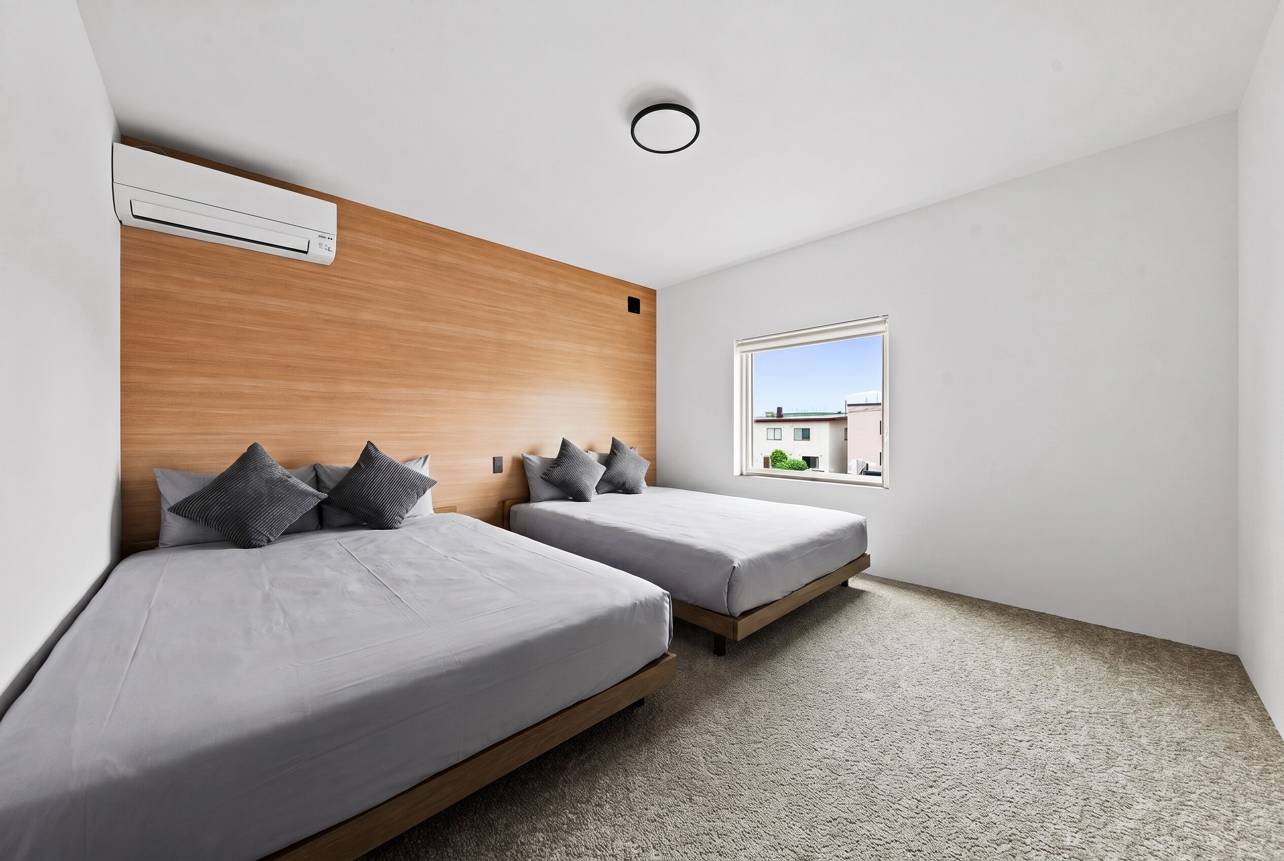 A modern bedroom with two beds, a wooden headboard, a window with a view, and a ceiling air conditioner.