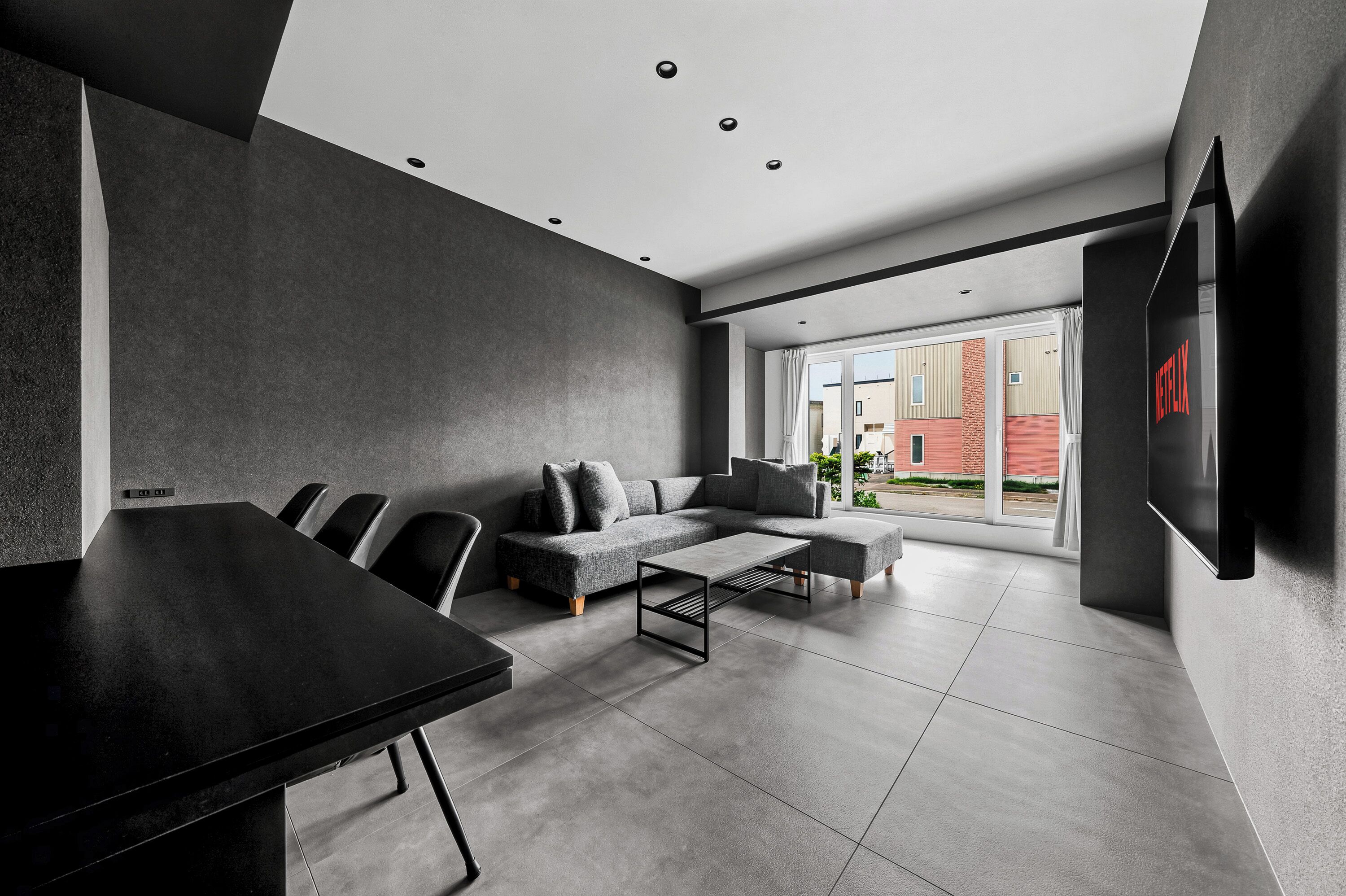 A modern living room with a sofa, coffee table, and chairs, a flat-screen TV mounted on the wall, and a large window offering a view of the outside.
