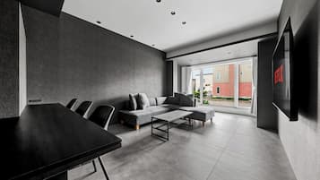 A modern living room with a sofa, coffee table, and chairs, a flat-screen TV mounted on the wall, and a large window offering a view of the outside.