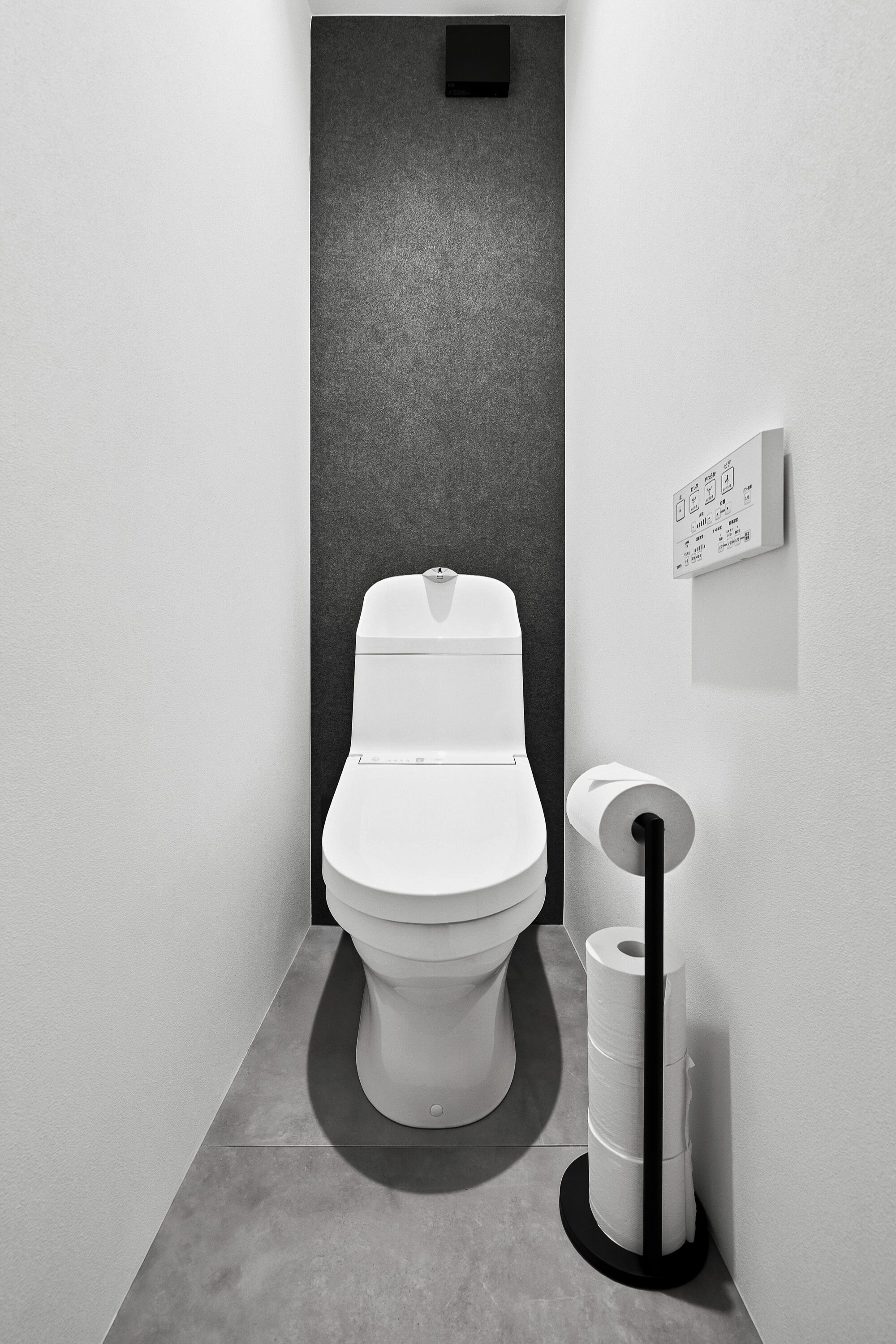 Un baño moderno y minimalista con inodoro blanco, pared de textura oscura y un dispensador de papel higiénico.