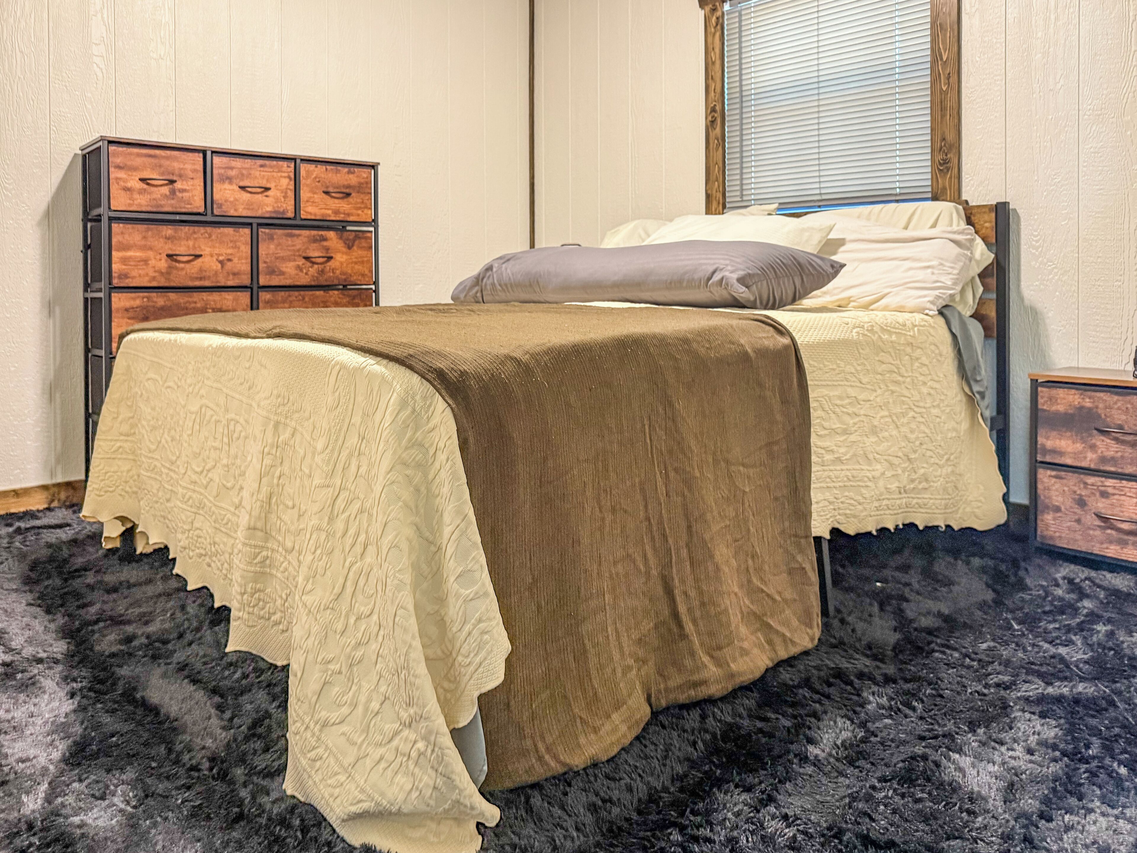 Una cama con colcha color beige y marrón, un aparador de madera con varios cajones, y una ventana con persianas.