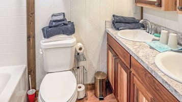 A bathroom with a toilet, two sinks, a trash can, and a wooden cabinet.