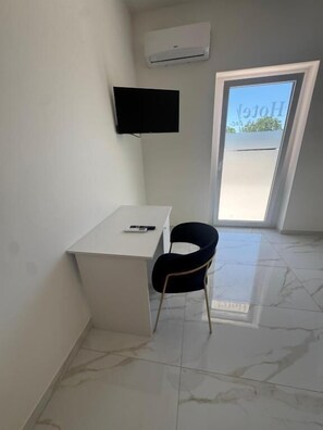 A room with a white desk, a black chair, a flat-screen TV mounted on the wall, and a sliding glass door leading to an outdoor area.