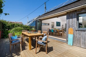 An outdoor deck with a wooden table and chairs, a surfboard leaning against a wall, and a clear blue sky. - An Mordros - sleeps 6 guests  in 3 bedrooms (Hayle)