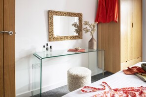 Ein modernes Schlafzimmer mit einem Glastischchen, einem Spiegel mit Steinrahmen, einem weißen Pouf und einem Bett mit roter Decke.
