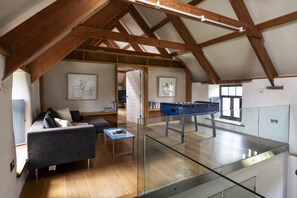 A spacious loft with a high ceiling, exposed wooden beams, a glass table, a sofa, and a foosball table. - Avallen Barn - sleeps 8 guests  in 4 bedrooms (Helford)