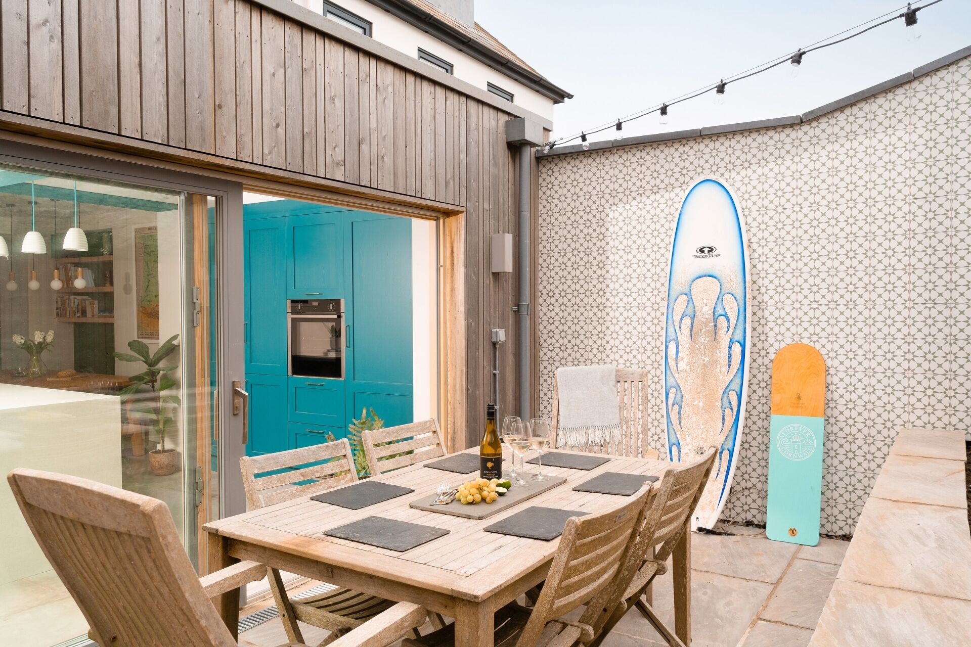 Un área de comedor al aire libre con una mesa y sillas de madera, una puerta azul y una tabla de surf apoyada en una pared con diseño.