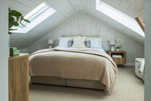 A bedroom with a sloped ceiling, a bed with a beige blanket, two bedside tables with lamps, and a plant. - Blue Haze - sleeps 6 guests  in 3 bedrooms (Hayle)