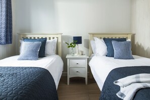 A hotel room with two beds, each with a white and blue patterned bedspread, a bedside table with a lamp, a vase with a plant, and a small drawer. - Blue Haze - sleeps 6 guests  in 3 bedrooms (Hayle)