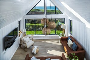 A spacious loft with a high ceiling, large windows offering ocean views, a cozy seating area with a sofa and armchair, and a hanging rattan light fixture. - Blue Haze - sleeps 6 guests  in 3 bedrooms (Hayle)