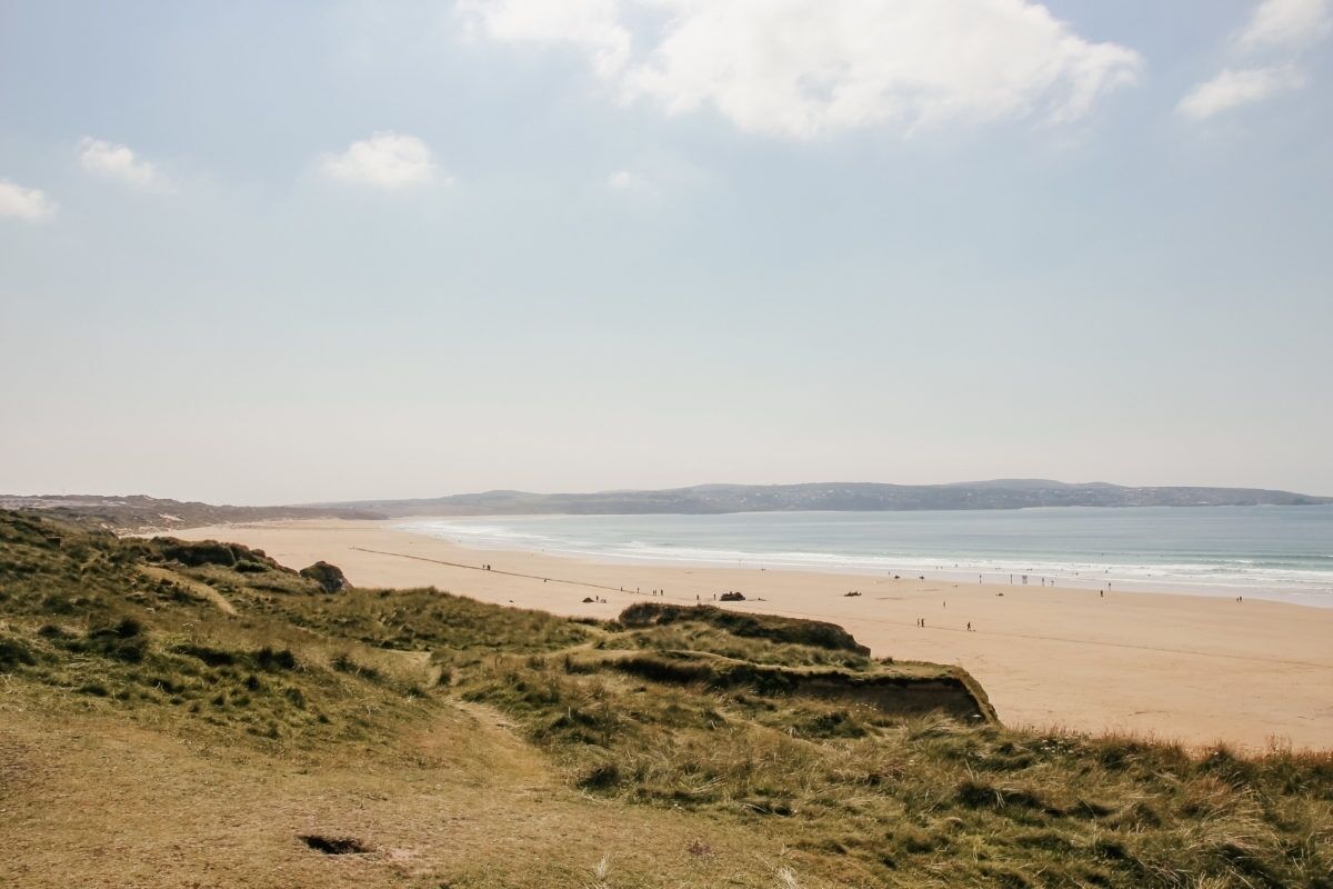 Une plage avec des dunes herbeuses au premier plan et une plage de sable où l’on aperçoit des personnes au loin.