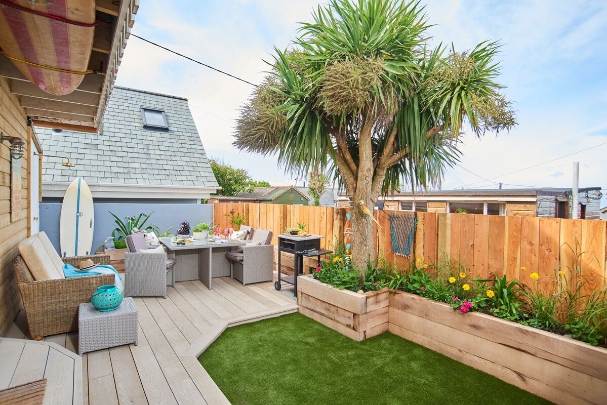 Ein Garten mit einer Holzterrasse, Gartenmöbeln, einer Palme und einem Blumenbeet.