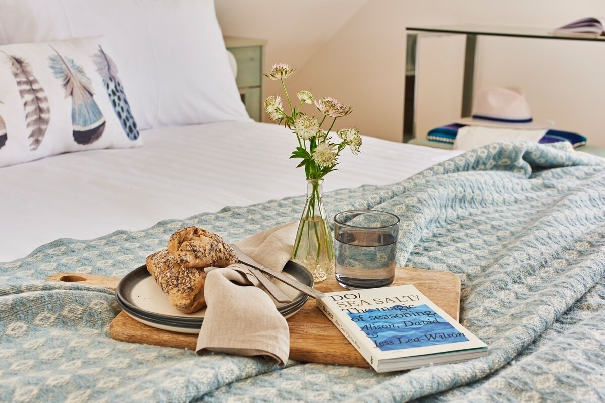 A neatly made bed with a blue patterned blanket, a pillow with feather designs, a book titled 'DO SEA SAINTS', a glass of water, and a plate with a croissant and a knife.