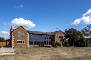 A two-story brick house with a large glass window wall on the ground floor, a chimney, and a fenced yard. - The Cottages Collection at Rusty Lane Farm (Seend)