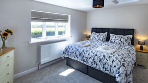A bedroom with a bed, bedside lamps, a radiator, a window with blinds, and a vase with flowers on a dresser. - The Cottages Collection at Rusty Lane Farm (Seend)