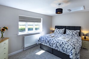 A bedroom with a bed, bedside lamps, a radiator, a window with blinds, and a vase with flowers on a dresser. - The Cottages Collection at Rusty Lane Farm (Seend)