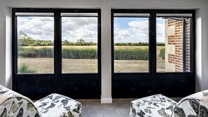 A room with two large windows offering a view of a field and a brick building. There are two patterned ottomans in the foreground. - The Cottages Collection at Rusty Lane Farm (Seend)