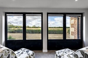 A room with two large windows offering a view of a field and a brick building. There are two patterned ottomans in the foreground. - The Cottages Collection at Rusty Lane Farm (Seend)