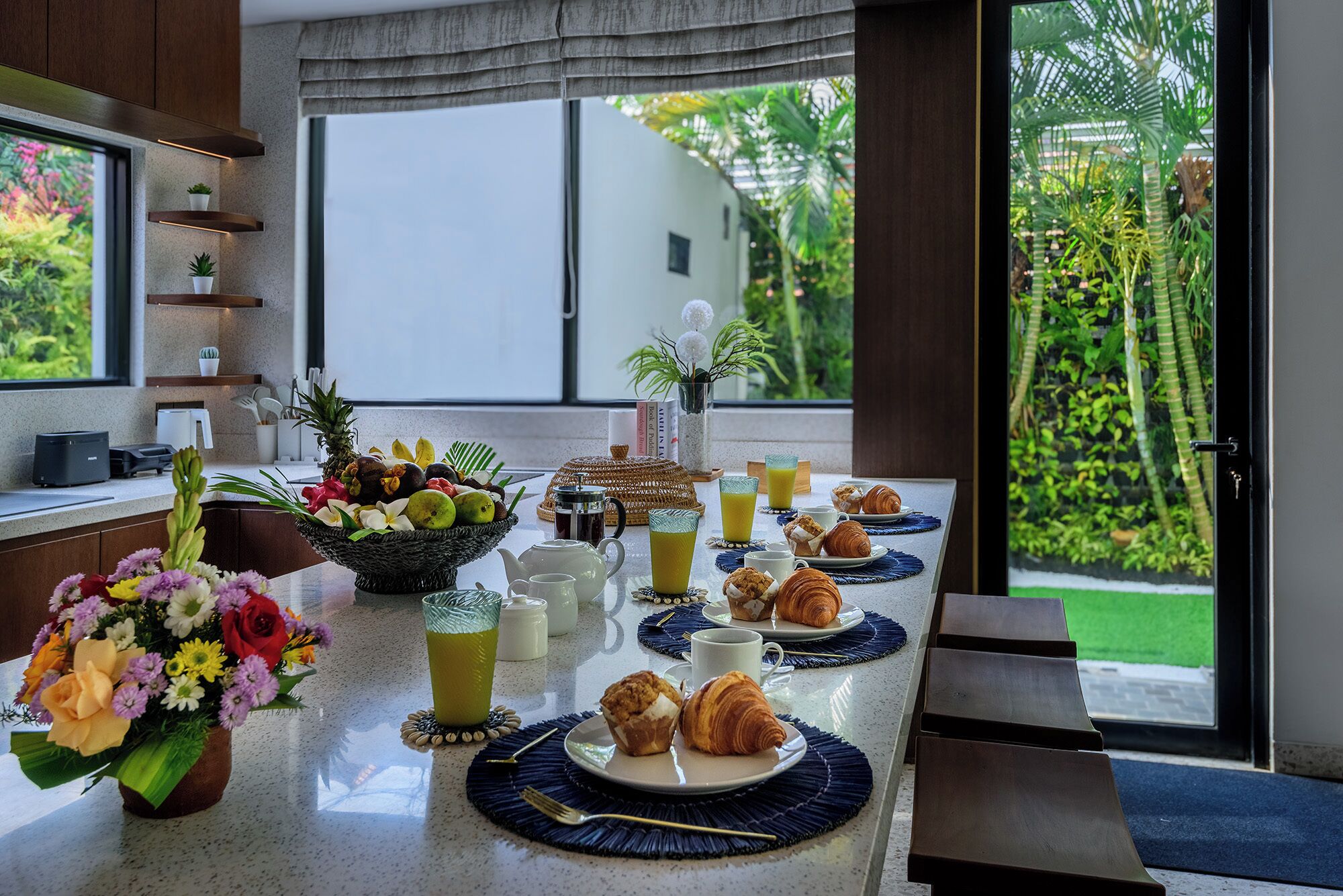 A breakfast setup with a fruit bowl, croissants, and juice glasses on a table with a view of a garden.