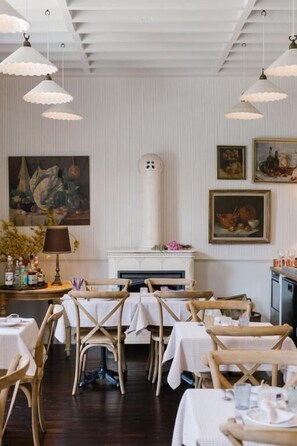 A dining area with tables set for guests, a fireplace, and wall-mounted artwork. - La Maison d'Hôtes (SILLY)