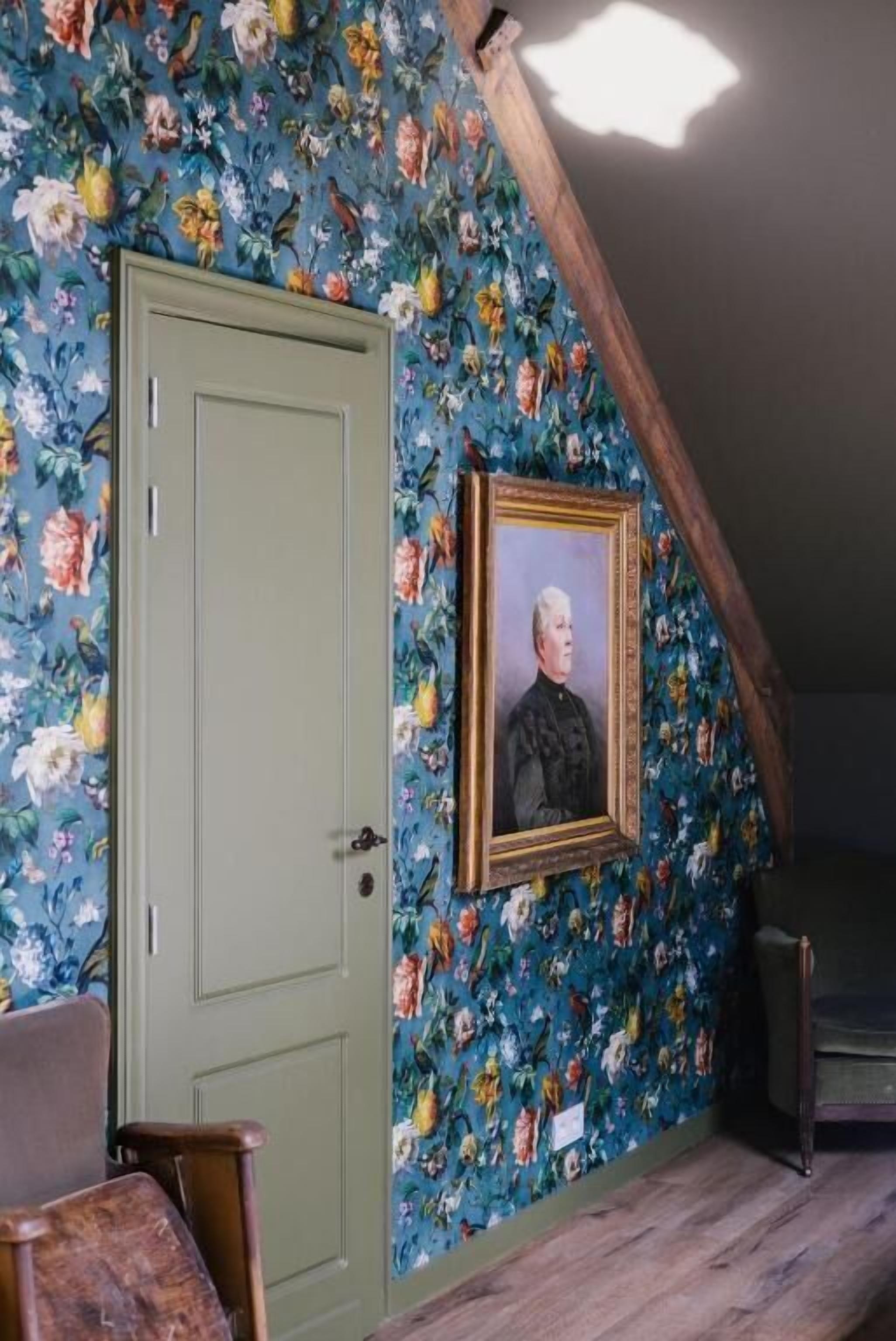 Een kamer met bloemenbehang, een ingelijst portret, een groene deur en een houten bank.