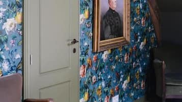 Een kamer met bloemenbehang, een ingelijst portret, een groene deur en een houten bank.