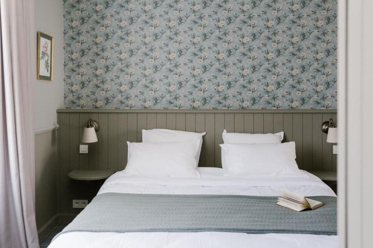 A bedroom with a bed, two bedside lamps, a nightstand, a framed picture, and a floral wallpaper.