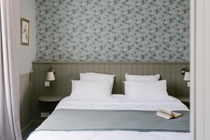 A bedroom with a bed, two bedside lamps, a nightstand, a framed picture, and a floral wallpaper. - La Maison d'Hôtes (SILLY)