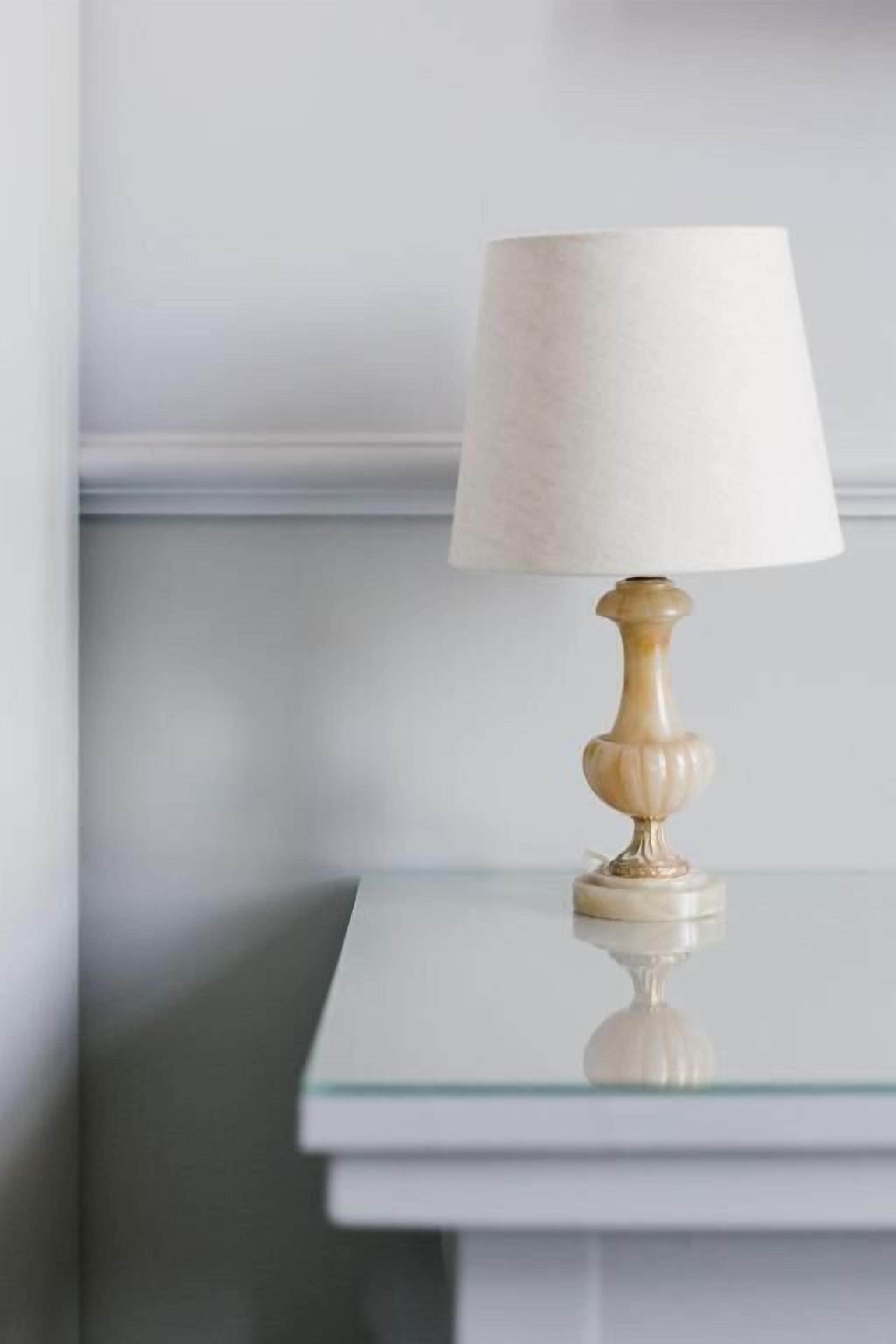 Eine Tischlampe mit weißem Lampenschirm auf einem Glastisch vor einer schlichten Wand.