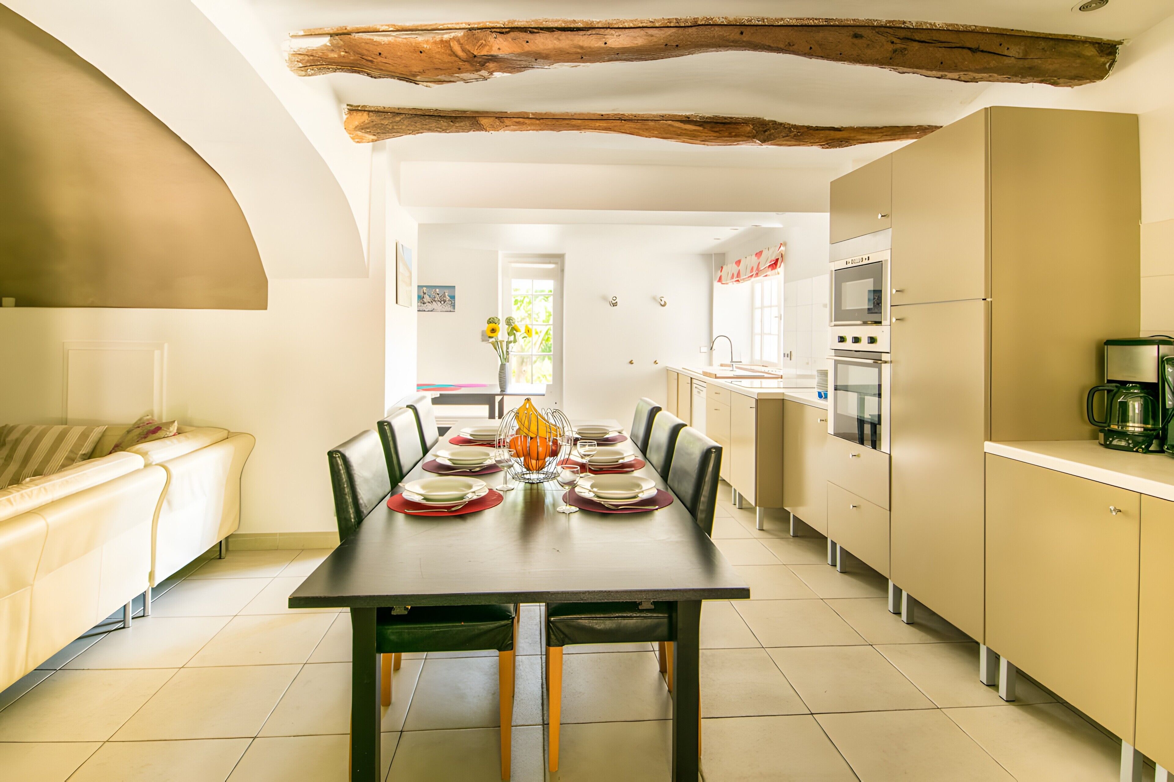 Un coin repas avec une longue table dressée pour quatre personnes, une cuisine équipée d’appareils modernes et un salon avec un canapé.