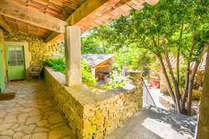 A stone-walled covered patio with a green door, outdoor seating, and a view of a house with a tiled roof. - Stenen Mas 'Le Mas Toupian'; 500m², Mediterranean Garden, Tennis, Pool, Pétanque, Air Con (Goudargues)