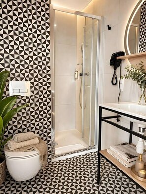 Ein Badezimmer mit Wand in geometrischem Muster, einer Duschkabine aus Glas, einer weißen Toilette, einem Waschbecken mit schwarzem Rahmen und einem Spiegel mit schwarzem Rahmen.