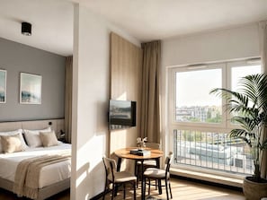 A hotel room with a bed, a TV mounted on the wall, a dining table with chairs, and a large window with a view of the city. - Old Town Heart - National Stadium Tower 87 (Warsaw)