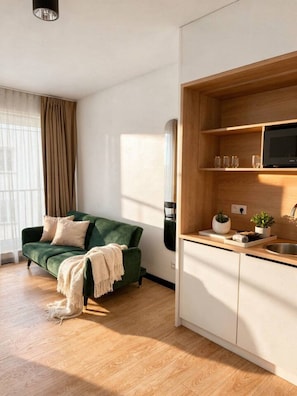 Un espace de vie moderne avec un canapé vert, un plancher en bois, un coin cuisine équipé d’un four à micro-ondes et une fenêtre avec des rideaux.