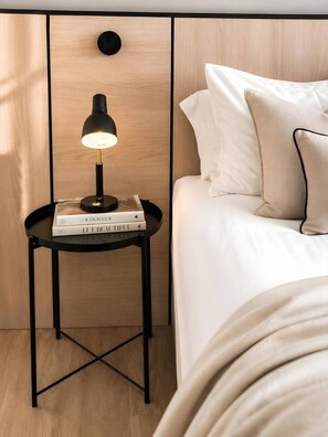 A neatly made bed with white bedding and a pillow, a small black side table with a lamp and books, and a wooden panel wall. - Old Town Heart - National Stadium Tower 22 (Warsaw)