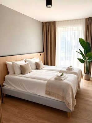 Ein Hotelzimmer mit zwei Betten, einem großen Fenster mit Vorhängen, einer Topfpflanze und einer Deckenleuchte.