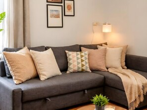 A sectional sofa with multiple cushions, a small plant on a side table, and wall-mounted lighting. - Old Town Heart - XXVII - Brzozowa (Warsaw)