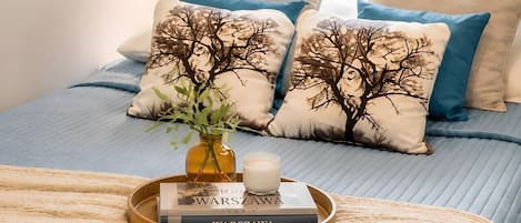 A bed with a blue quilt, two pillows with tree designs, a bedside table with a tray holding books and a candle, and a vase with greenery.
